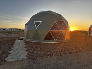 A geodesic dome structure with a triangular window and a door, situated on a gravel path with a sunset in the background. - Amazing Dome with the Greatest Desert Views Ever in Willcox, Arizona (Wilcox)