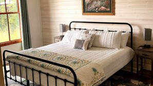 Un dormitorio con cama, mesita de noche, lámpara, ventana con cortinas e imagen enmarcada en la pared.
