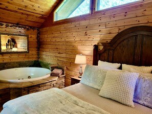 Quarto de cabana de tronco com interior de madeira, uma banheira grande, cama com roupa de cama branca e janela com vista para árvores.