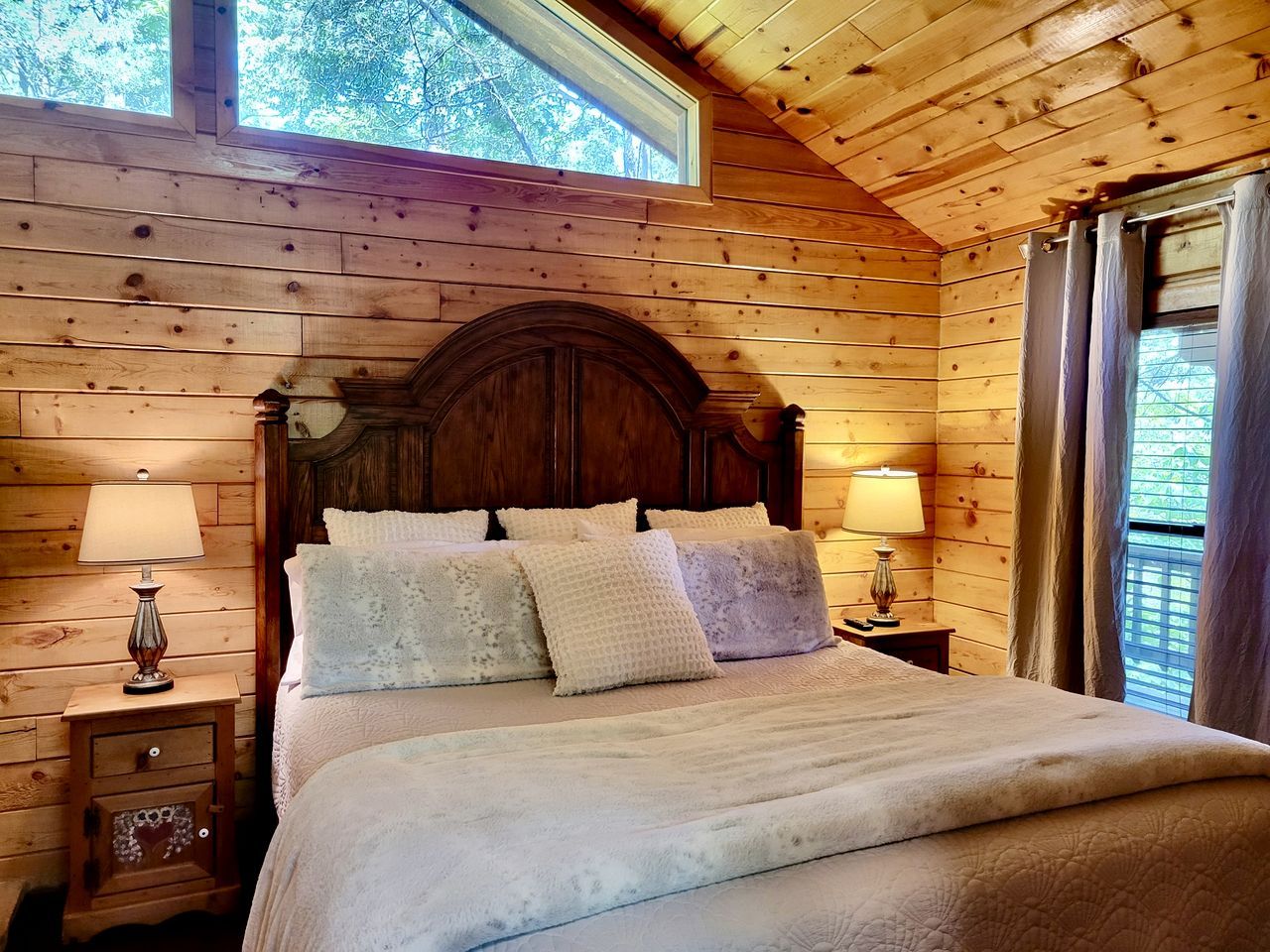 Ein Schlafzimmer einer Holzhütte mit einem großen Bett, zwei Nachttischen mit Lampen und einem Fenster mit Vorhängen.