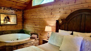 A log cabin room with a wooden interior, a large bathtub, a bed with white bedding, and a window with a view of trees. - Rustic Cabin with Indoor Bath Spa and Deck in Arkansas (Eureka Springs)