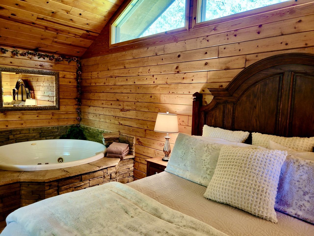 Una habitación tipo cabaña de madera con interior de madera, una bañera grande, una cama con ropa blanca y un ventanal con vistas a los árboles.