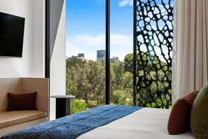 A modern hotel room with a large window, a bed with a blue blanket, a wooden bench, and a flat-screen TV mounted on the wall. - Hackney Hotel (Hackney)