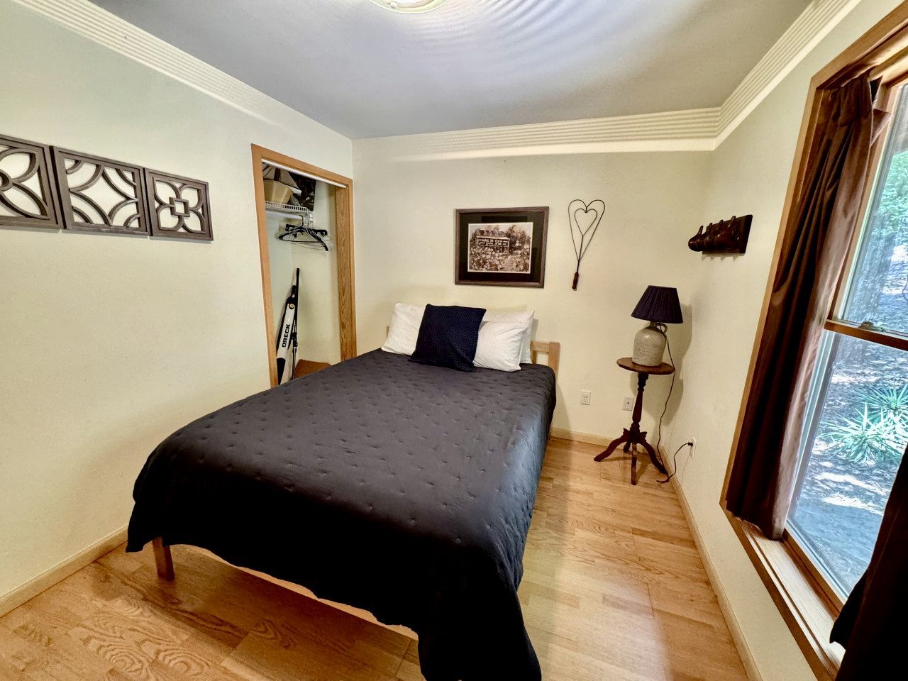 A bedroom with a bed, a small table with a lamp, a framed picture, and a window with curtains.