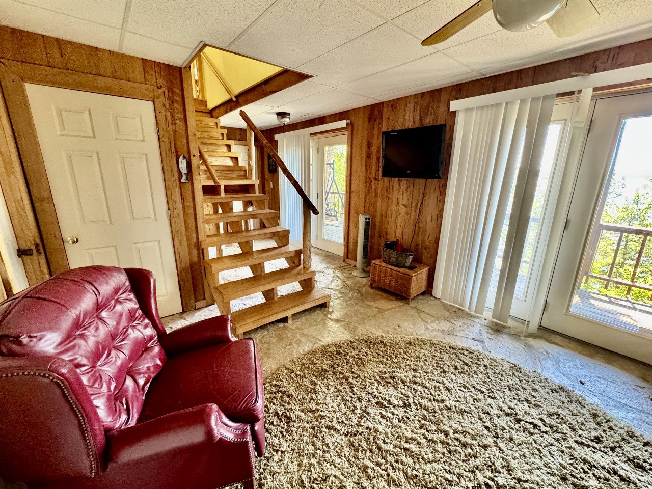 A living room with a red leather sofa, a wooden staircase, a flat-screen TV, and a large window with curtains.