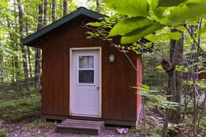 Een klein houten vakantiehuisje met een witte deur en een raam, omgeven door een bos.