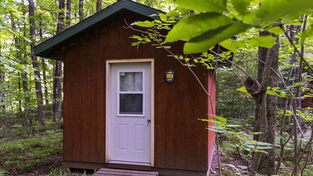 Une petite cabane en bois, avec une porte blanche et une fenêtre, entourée d’une forêt.
