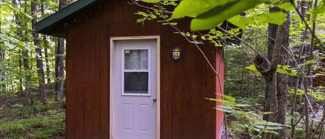 Una pequeña cabaña de madera con una puerta blanca y una ventana, rodeada por un bosque.