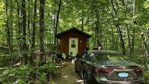 Une voiture est garée dans une zone boisée, avec une petite cabane en arrière-plan.