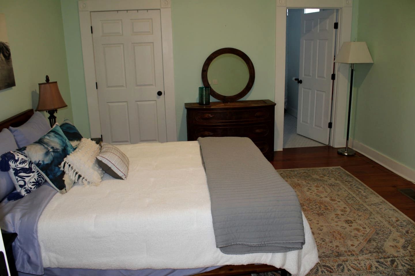 Une chambre à coucher comprenant un lit, une commode, un miroir, une lampe et une porte donnant sur une autre pièce.