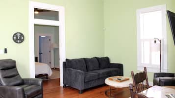 Un salon avec un canapé gris, une table à manger en bois, un fauteuil en cuir noir et une chaise en bois avec un dossier en osier tressé.