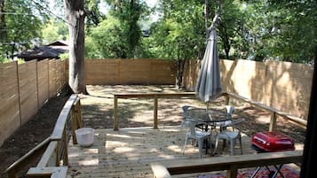 Une terrasse en bois avec une table et des chaises, un parasol suspendu et un objet rouge posé sur la table.