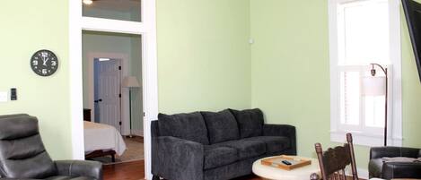 Una sala de estar con un sofá gris, una mesa de comedor de madera, un sillón de cuero negro y una silla de madera con respaldo tejido.