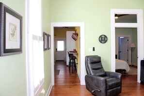 Un pasillo con cuadros enmarcados, un sillón de cuero, una zona de comedor con mesa y sillas, y un reloj en la pared.