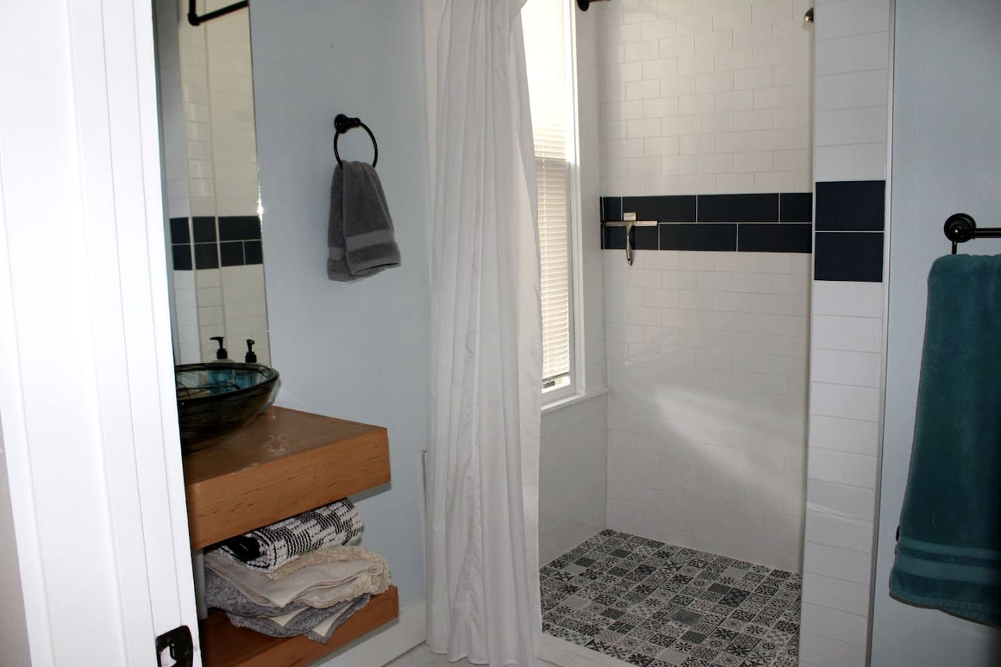 A bathroom with a wooden vanity, a glass bowl sink, a towel rack with a towel, a shower with black and white tiles, and a patterned floor.