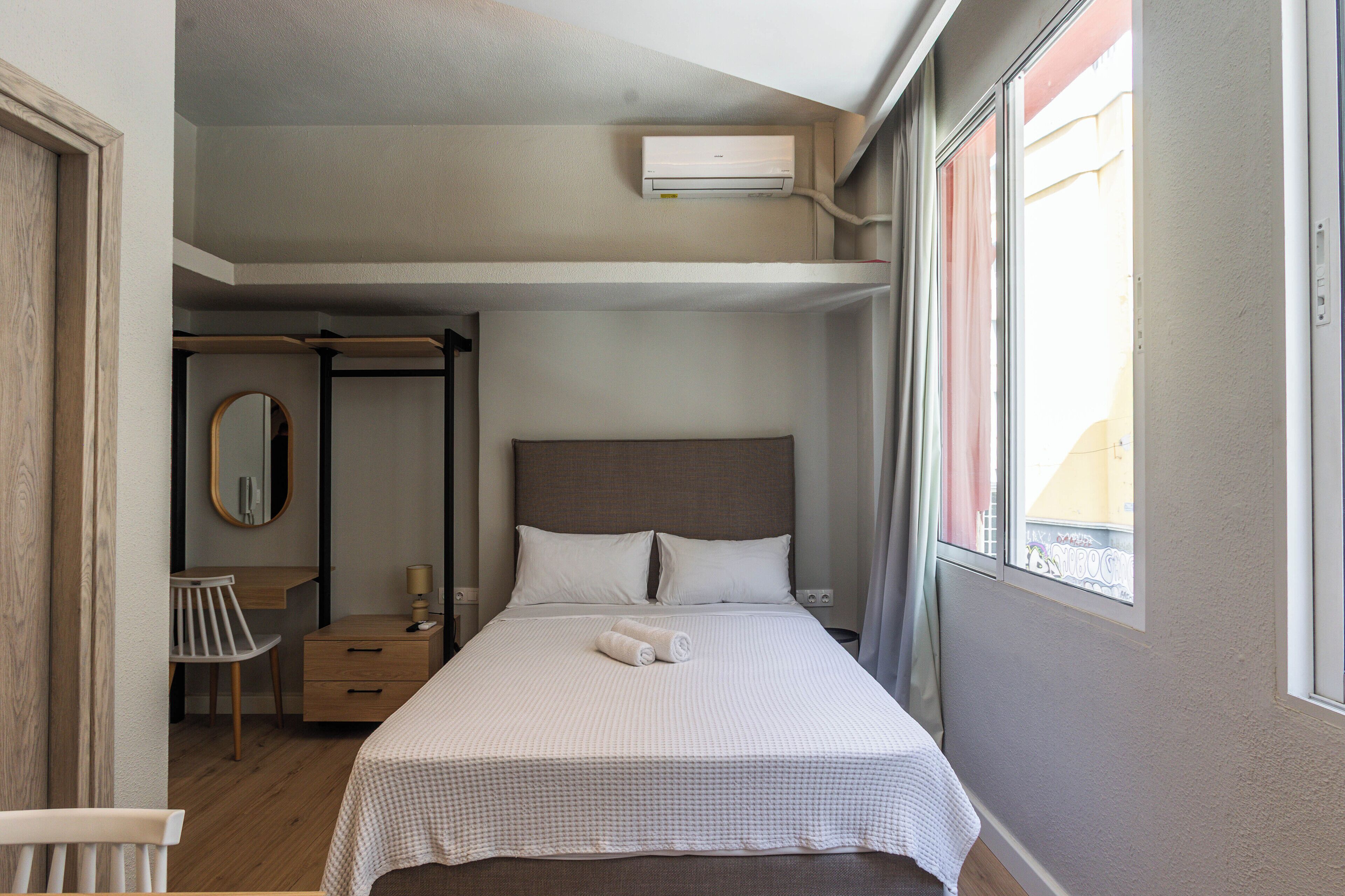 Quarto de hotel com cama, escrivaninha, cadeira, espelho e janela com cortinas.