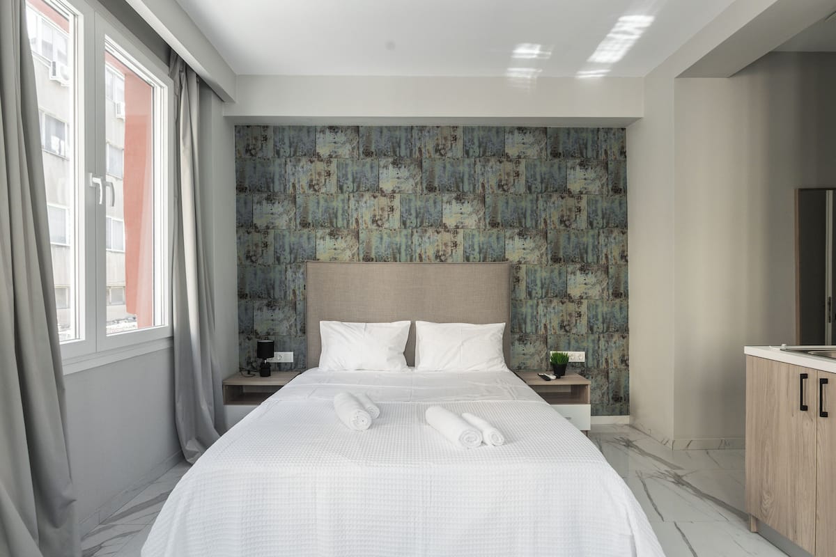 A modern hotel room with a large bed, a headboard with a textured wall, a bedside table with a lamp, and a view of the outside through a window.