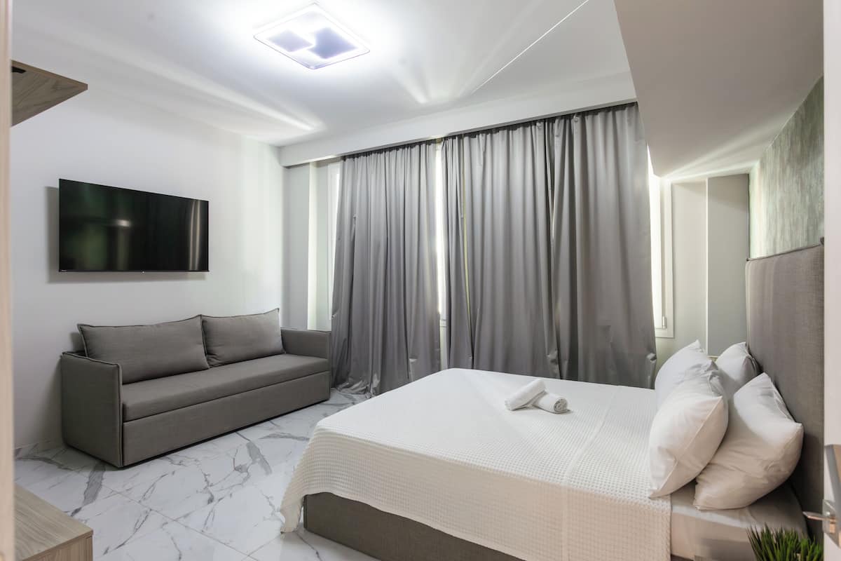 A modern hotel room with a large bed, a sofa, a flat-screen TV, and a large window with curtains.