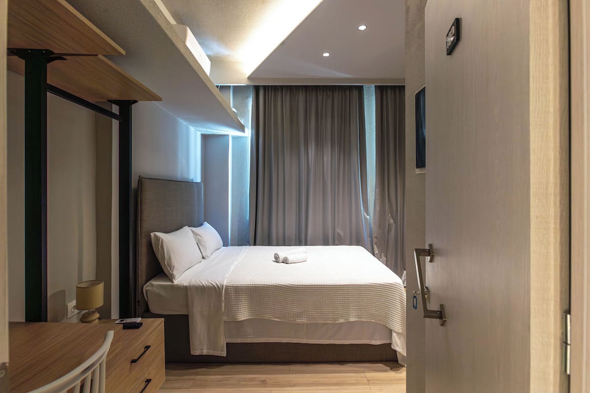 A modern hotel room with a neatly made bed, a wooden desk with a lamp, and a large window with curtains.