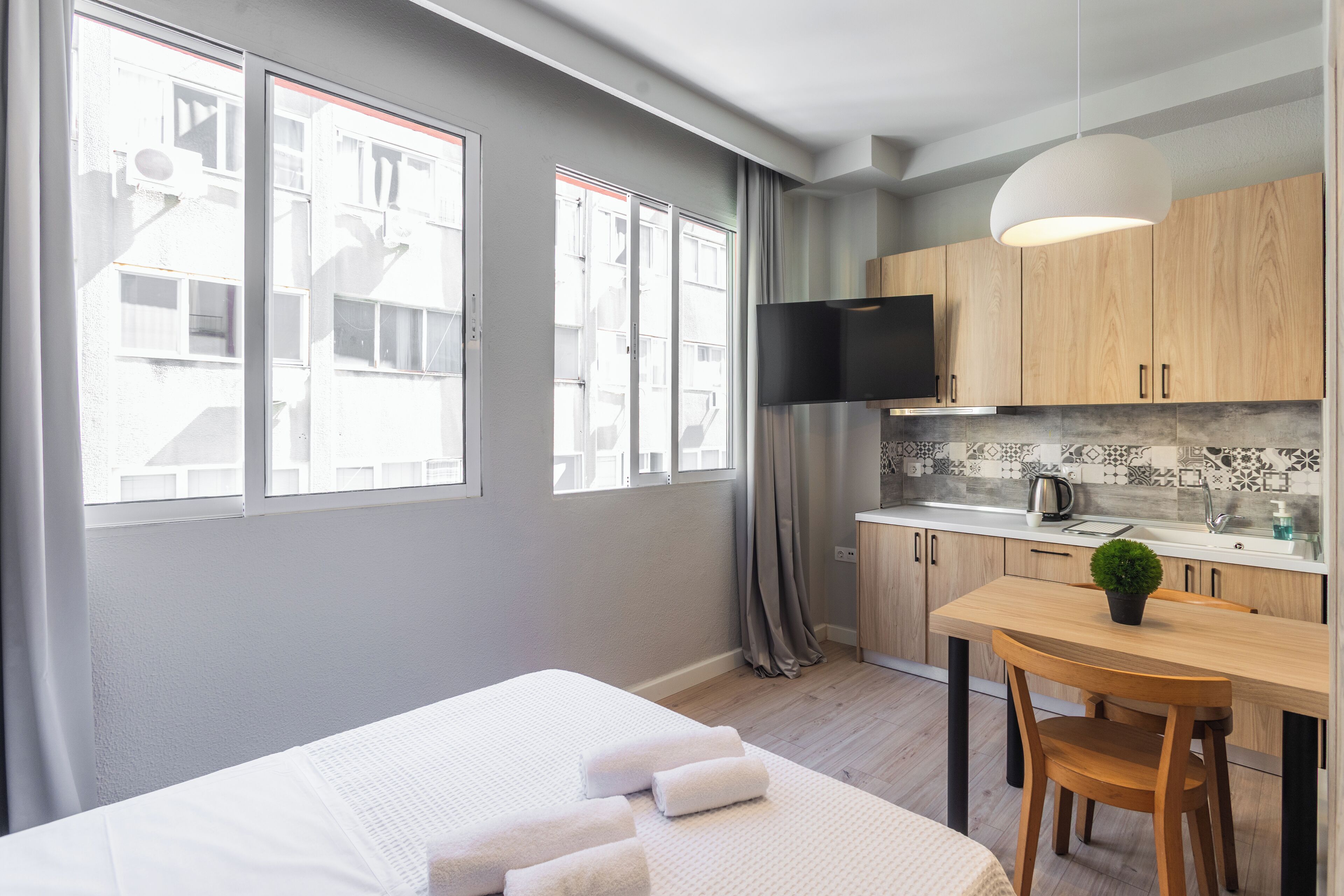 A modern hotel room with a bed, a dining table with chairs, a kitchen area with a TV, and large windows with curtains.