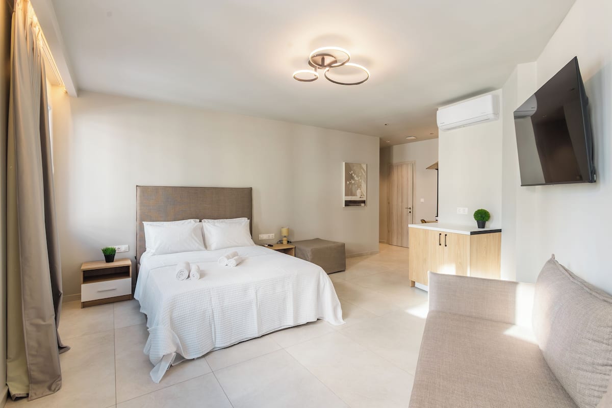 A modern hotel room with a large bed, bedside tables, a TV mounted on the wall, and a kitchenette area.
