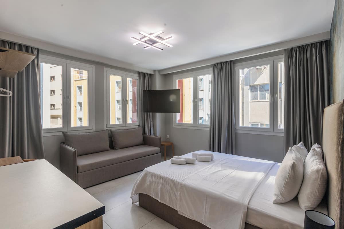 A modern hotel room with a bed, a sofa, a TV, and large windows.