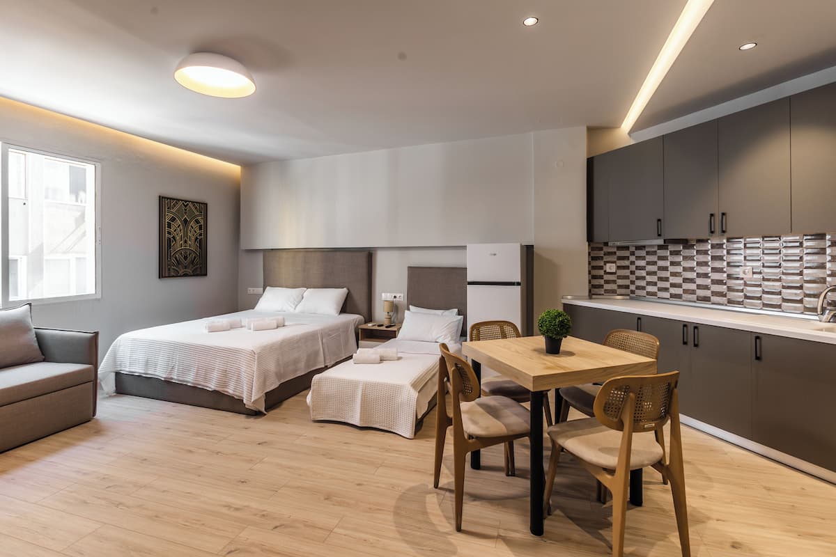 A modern hotel room with two beds, a dining area, and a kitchenette.