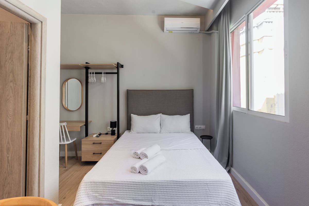 A modern hotel room with a neatly made bed, a desk with a chair, a mirror, and a window with curtains.