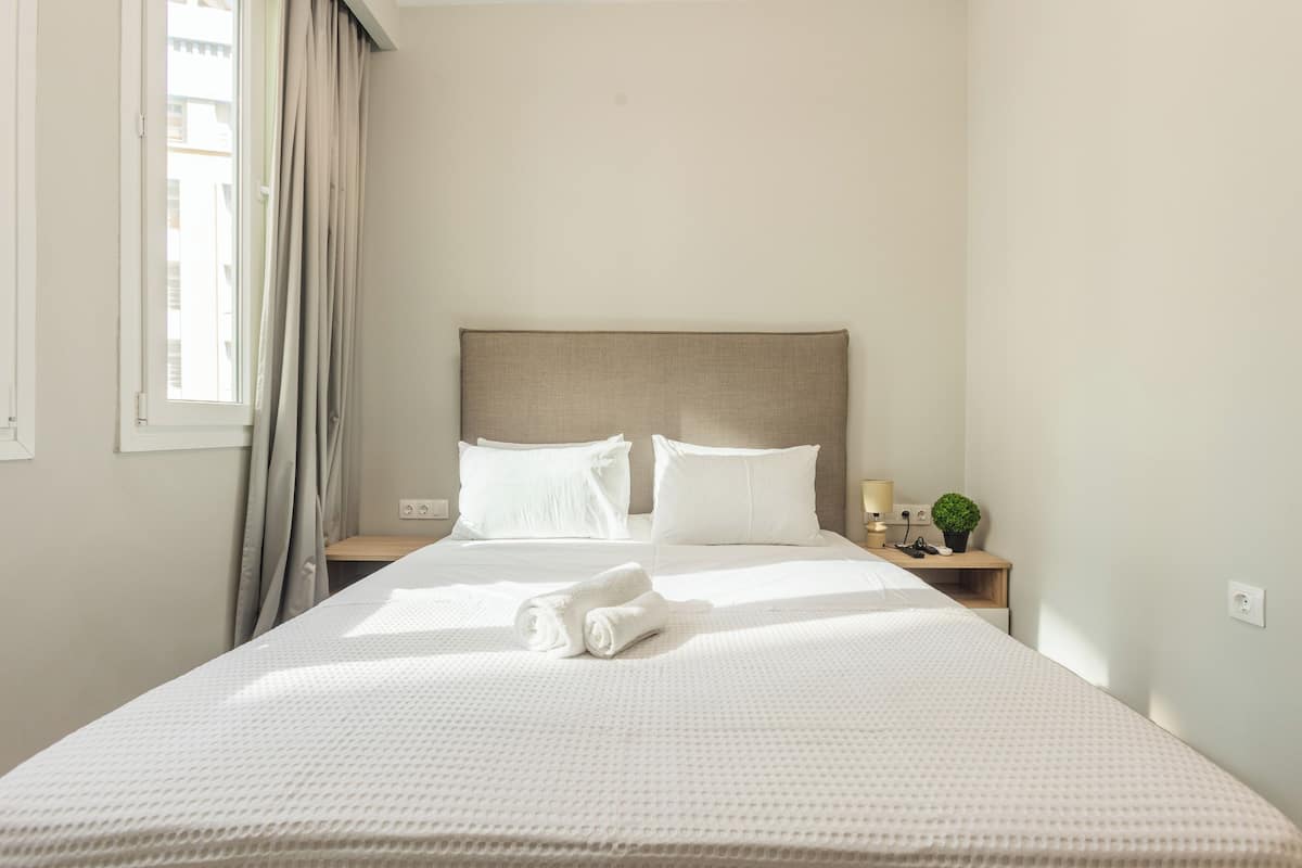 A neatly made bed with white linens, a headboard, and a small bedside table with a lamp and a plant.