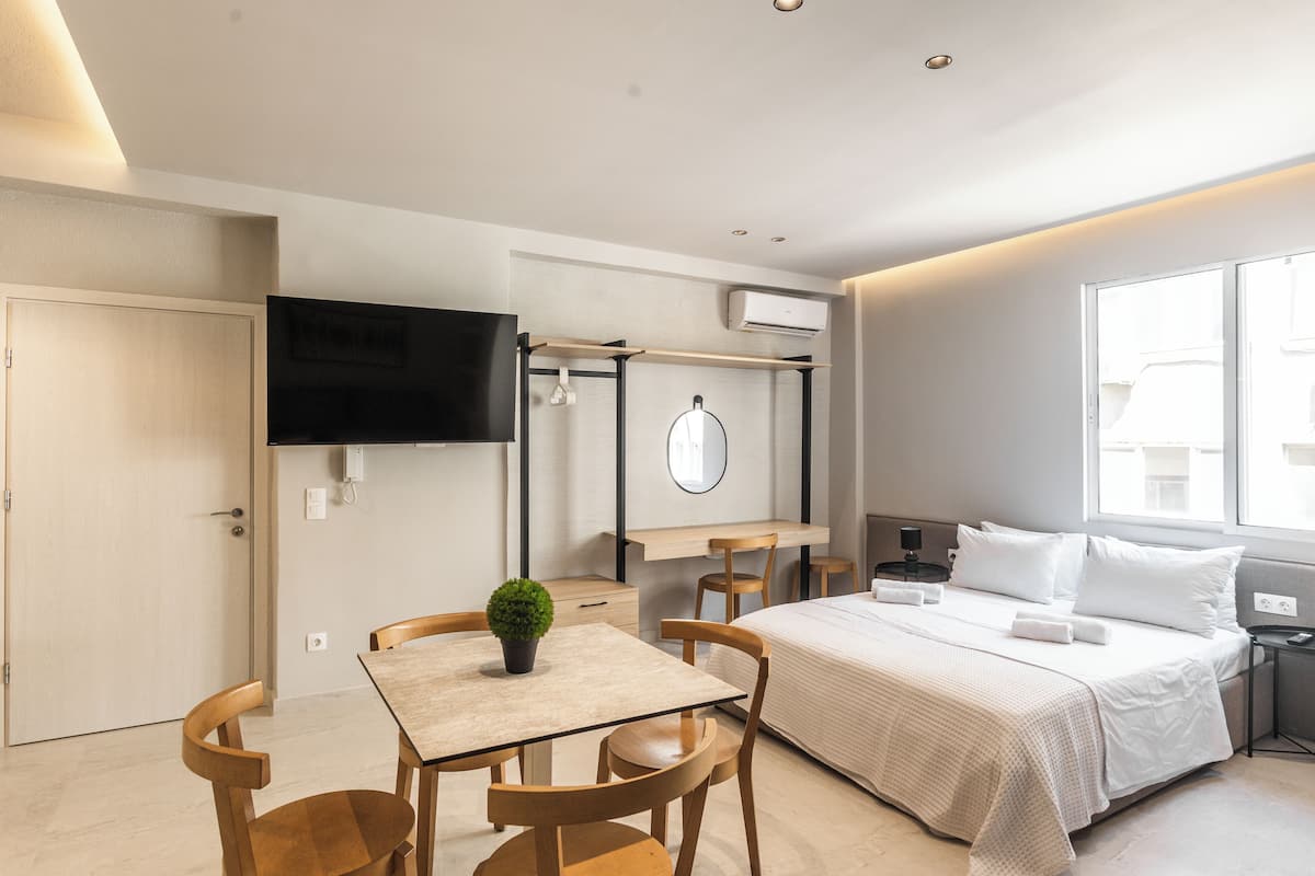 A modern hotel room with a bed, bedside table, TV, and a small dining area with chairs and a table.