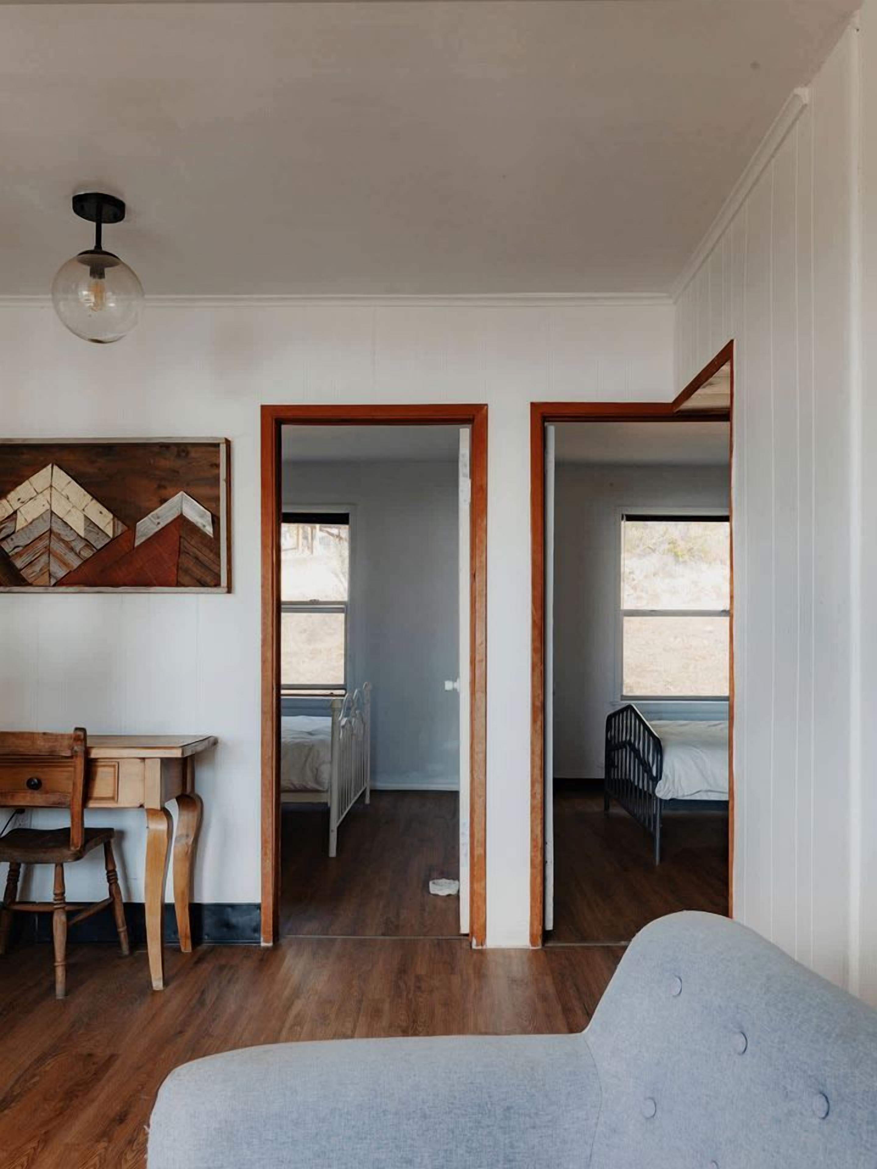 A room with a wooden desk, a gray sofa, and a bed with a black headboard. There are two doorways leading to other rooms.