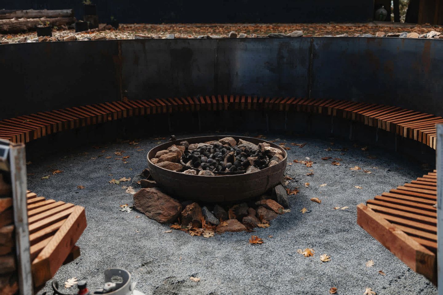 A circular fire pit with logs and rocks, surrounded by a concrete border with wooden steps.