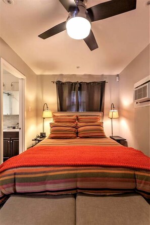 Un dormitorio con cama, ventilador de techo, dos lámparas de noche, ventana con cortinas y aire acondicionado de pared.