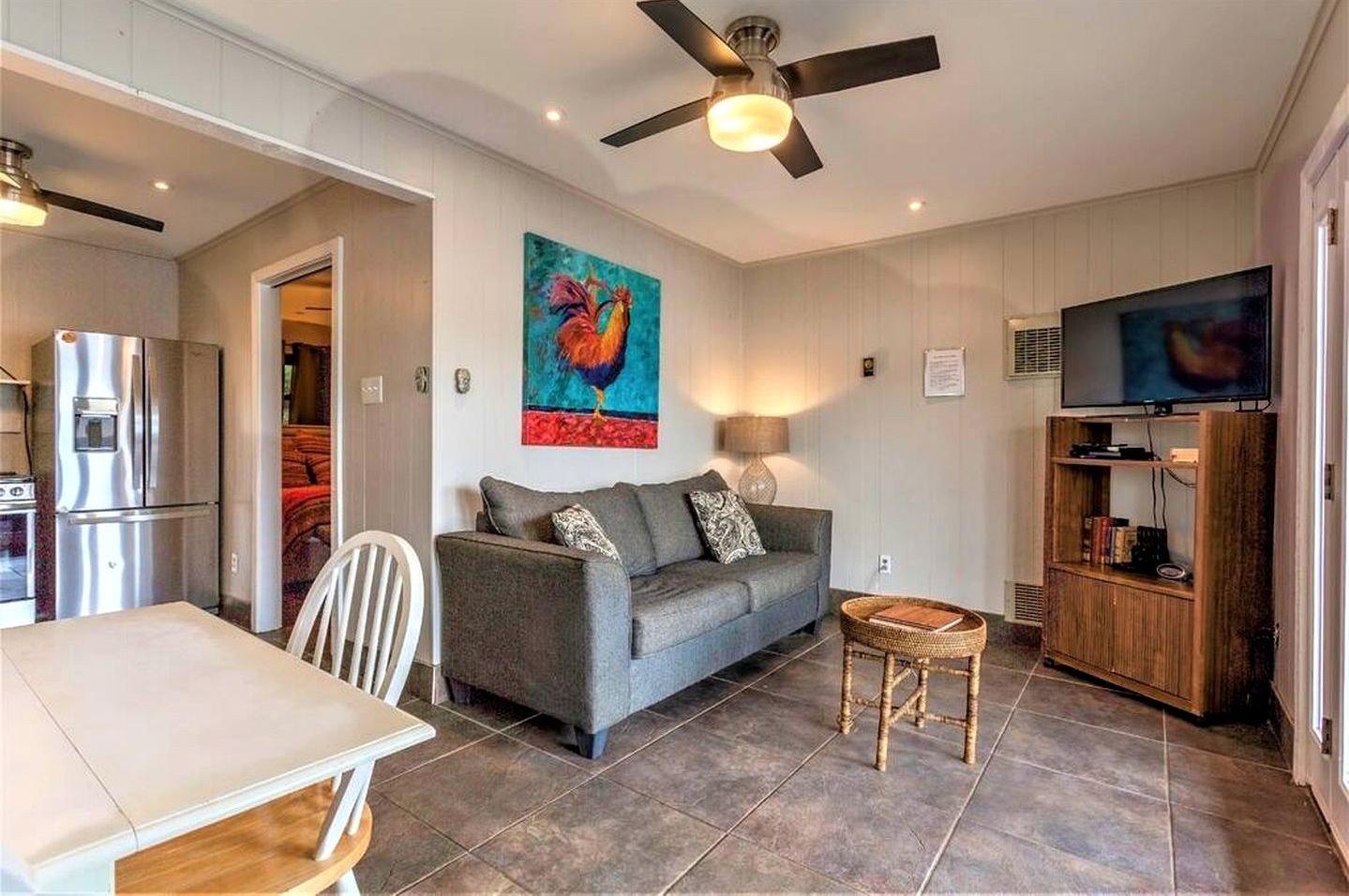 A compact living space with a kitchenette, a sofa, a dining table, a ceiling fan, a television, and a painting.