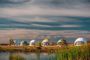 Une rangée de structures en forme de dômes géodésiques, situées au bord d’un plan d’eau, sous un ciel nuageux.