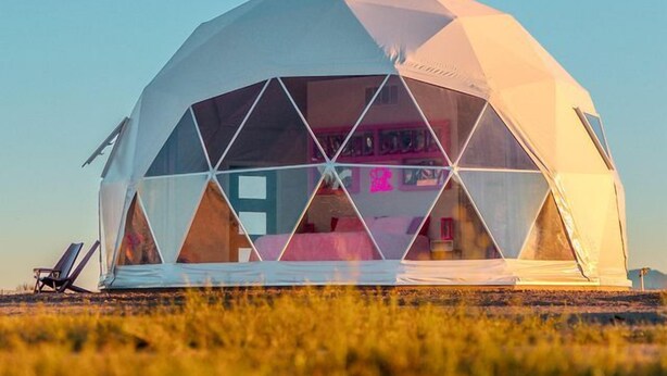 A geodesic dome structure with a transparent interior, a bed, and a chair outside.