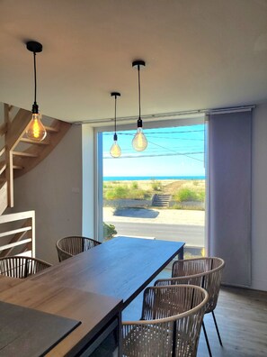 Un comedor moderno con una mesa y sillas de madera, lámparas colgantes y vistas a la playa a través de grandes ventanales.