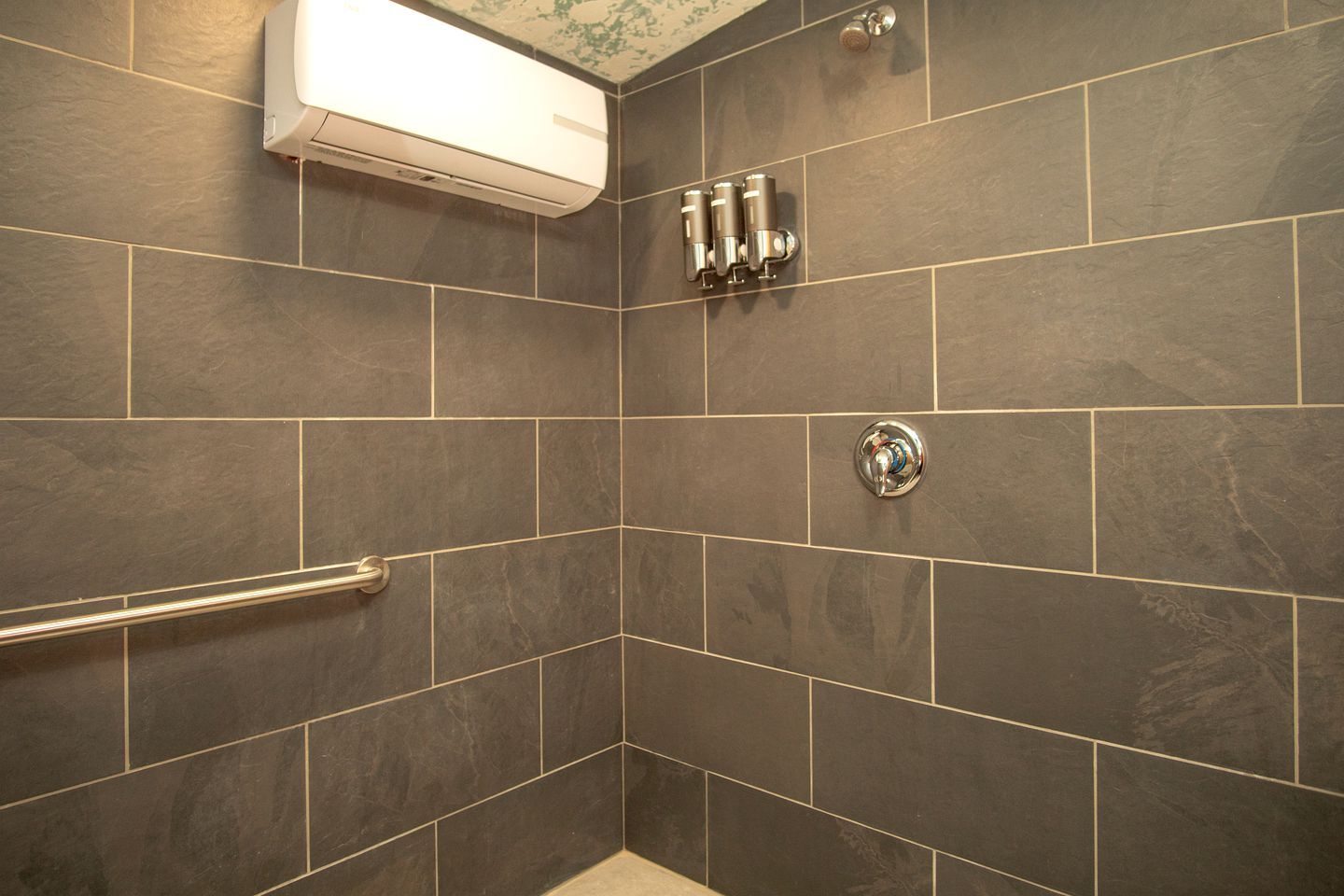 A shower with a handrail, a wall-mounted air conditioning unit, and a corner tile design.