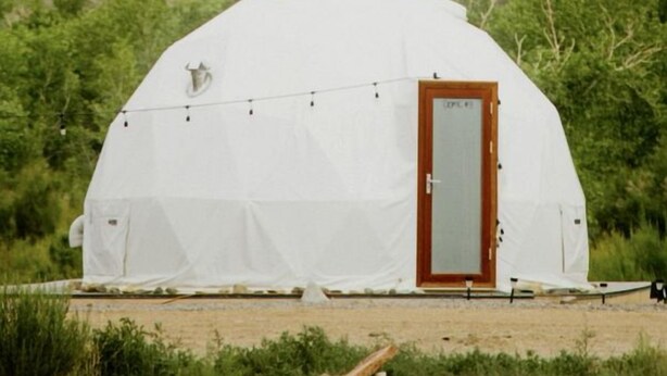 Una tienda de forma abovedada con puerta de madera, rodeada de vegetación y un risco rocoso al fondo.