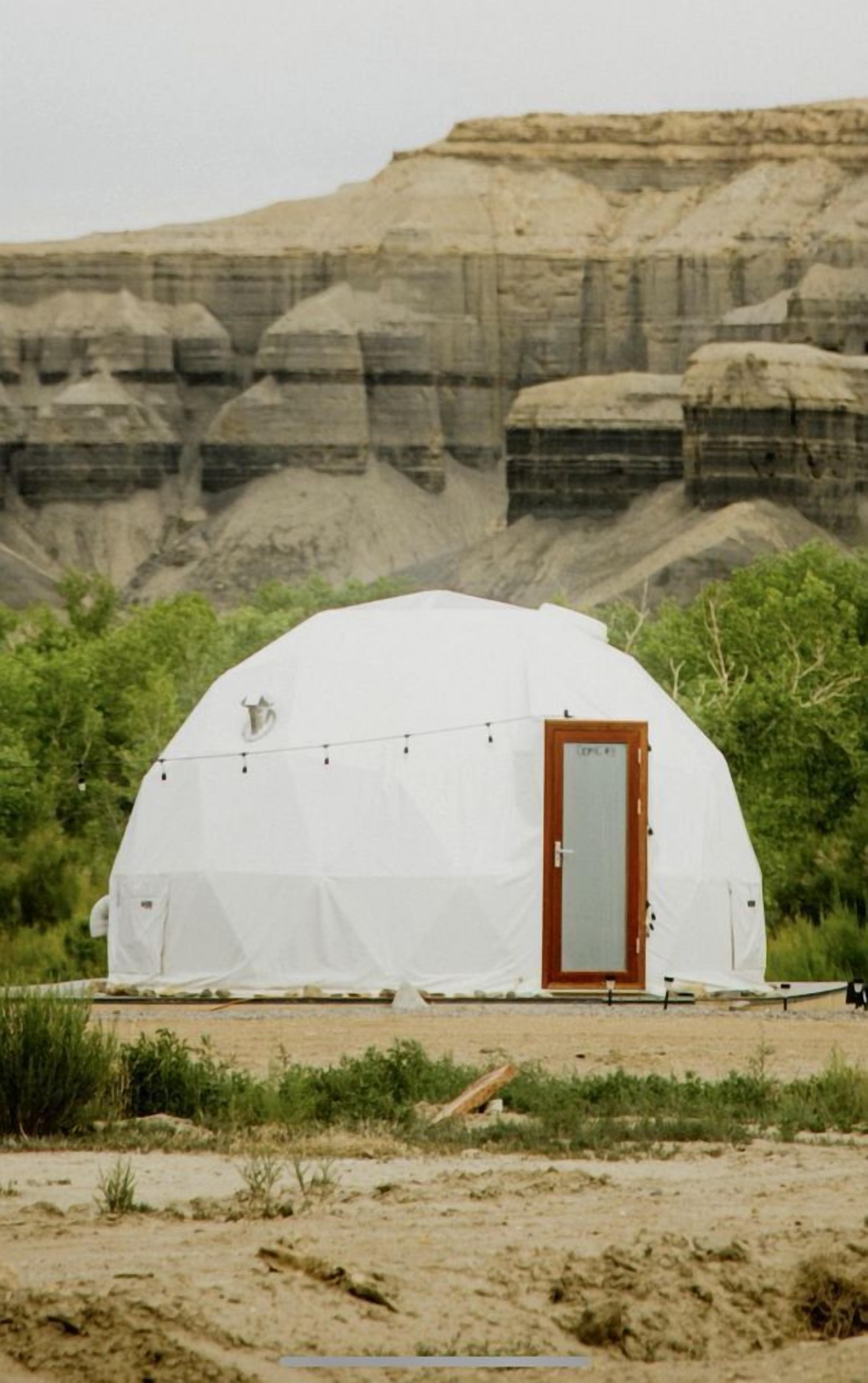 A dome-shaped tent with a wooden door, surrounded by greenery and a rocky cliff in the background.