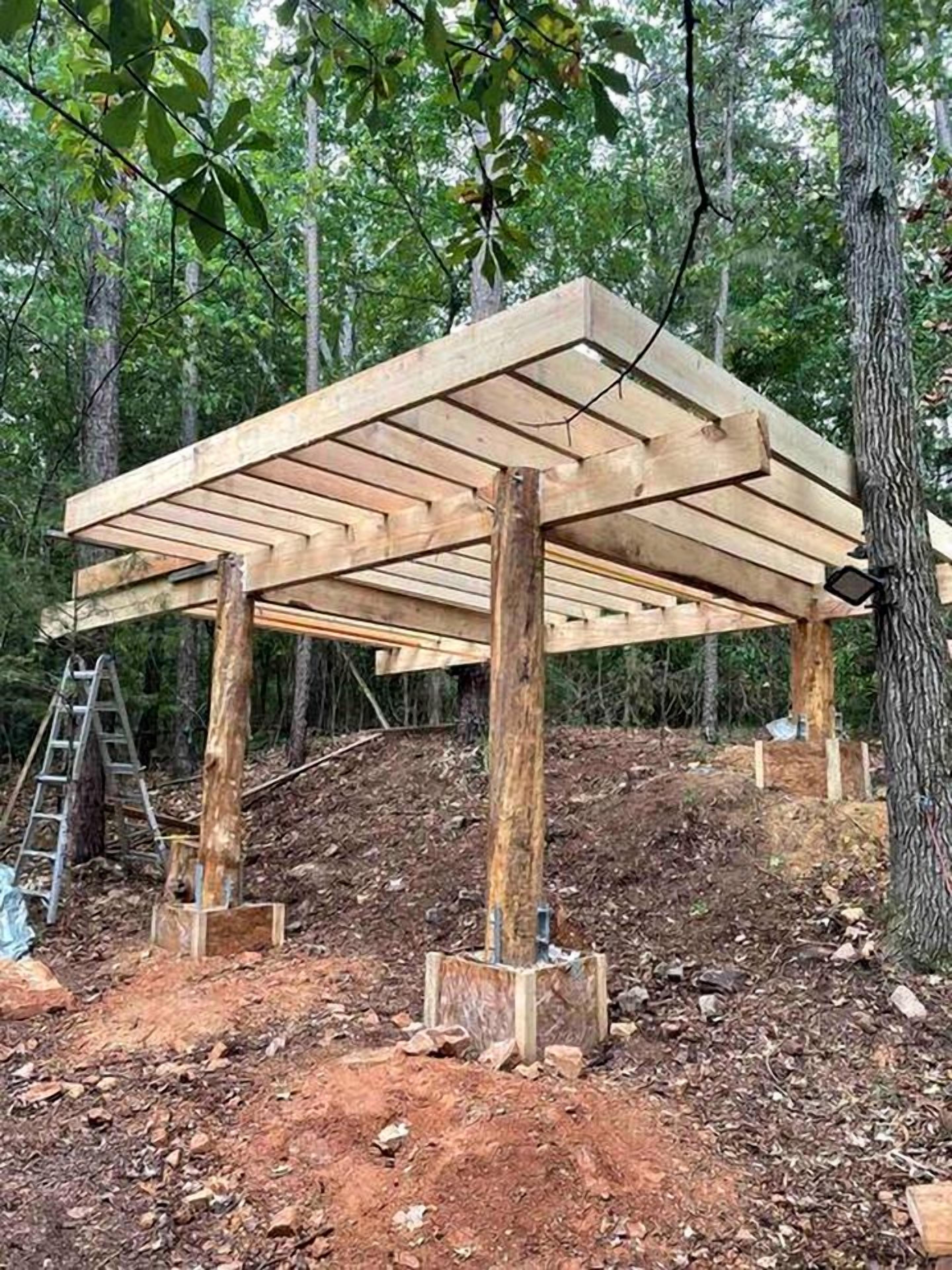 A wooden structure with a roof supported by beams, surrounded by trees.