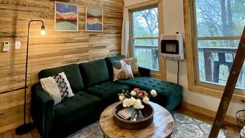 A cozy cabin interior with a wooden ceiling, a green sofa, a round coffee table with flowers, and large windows.