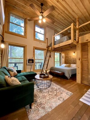 A cozy cabin interior with a loft bed, a green sofa, a round coffee table, and large windows. - Luminous Peaceful Treehouse with Incredible Forest Views and Fire Pit in Arkansas (Boles)