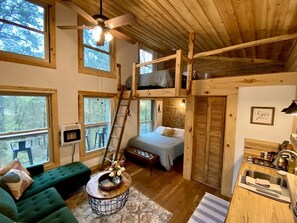 Interior acollidor d'una cabana amb un llit d'altell, un sofà verd, una taula de dinar de fusta i una vista del bosc a través de grans finestres.