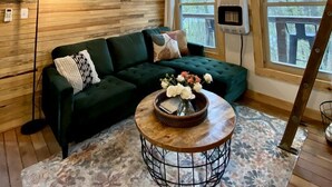 A cozy cabin interior with a wooden ceiling, a green sofa, a round coffee table with flowers, and large windows. - Luminous Peaceful Treehouse with Incredible Forest Views and Fire Pit in Arkansas (Boles)