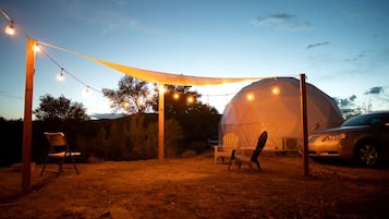 A geodesic dome tent with string lights, two chairs, and a car parked outside.