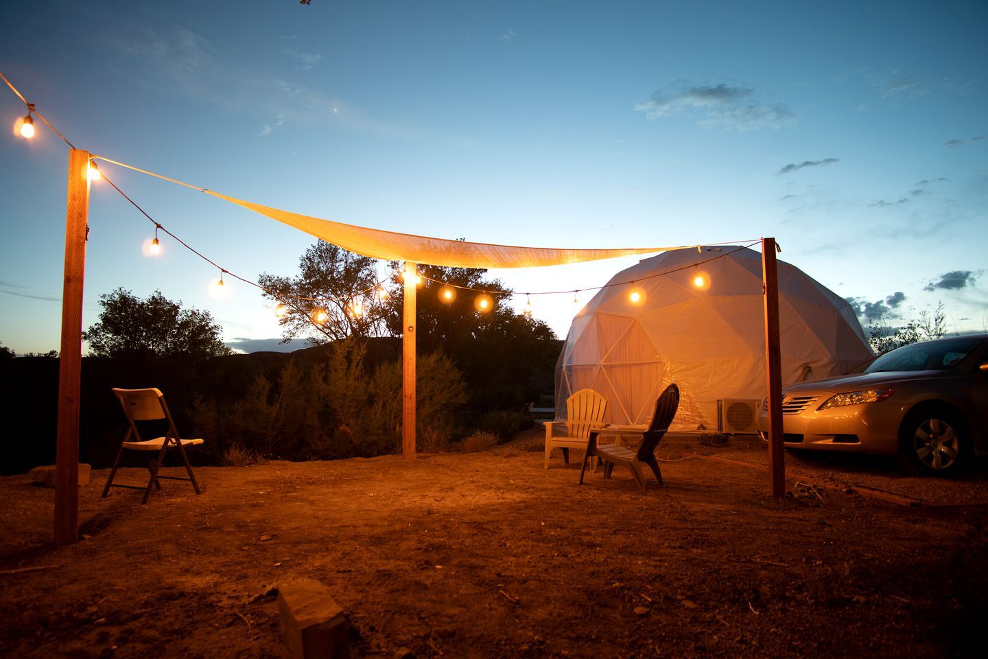 A geodesic dome tent with string lights, two chairs, and a car parked outside.