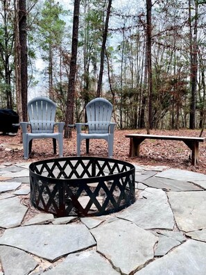 Un foyer en pierre, entouré de chaises Adirondack et d’un banc, dans un cadre forestier.