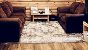 Una habitación acogedora con paredes de madera, un ventilador de techo, un televisor de pantalla plana, dos sofás de cuero marrón, una pequeña mesa de centro y cuadros enmarcados en la pared.