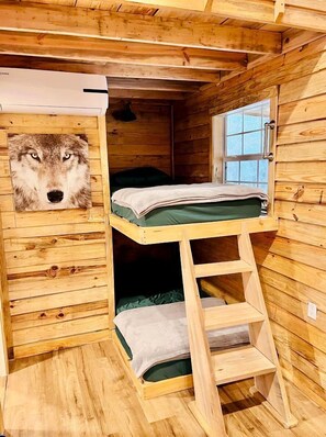 Una habitación de cabaña de madera con una cama individual superpuesta, una ventana y un aire acondicionado instalado en la pared.